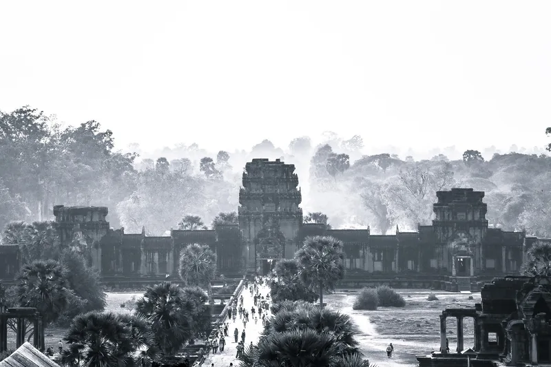 Silhouetted towers of Angkor Wat rise above mist and trees at dawn, with visitors walking the causeway in Siem Reap.