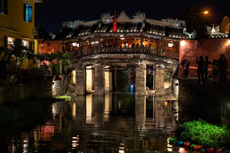 The Japanese Covered Bridge glows at night with tourists crossing above its reflection in Hoi An's canal.