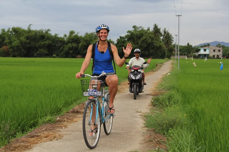 A woman waves while cycling along a rural path through green rice paddies near Hoi An.