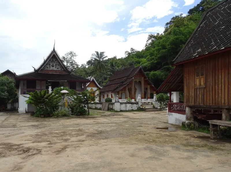 Traditional Lao temple buildings with tiered roofs surround a dirt courtyard at a monastery in Luang Prabang.