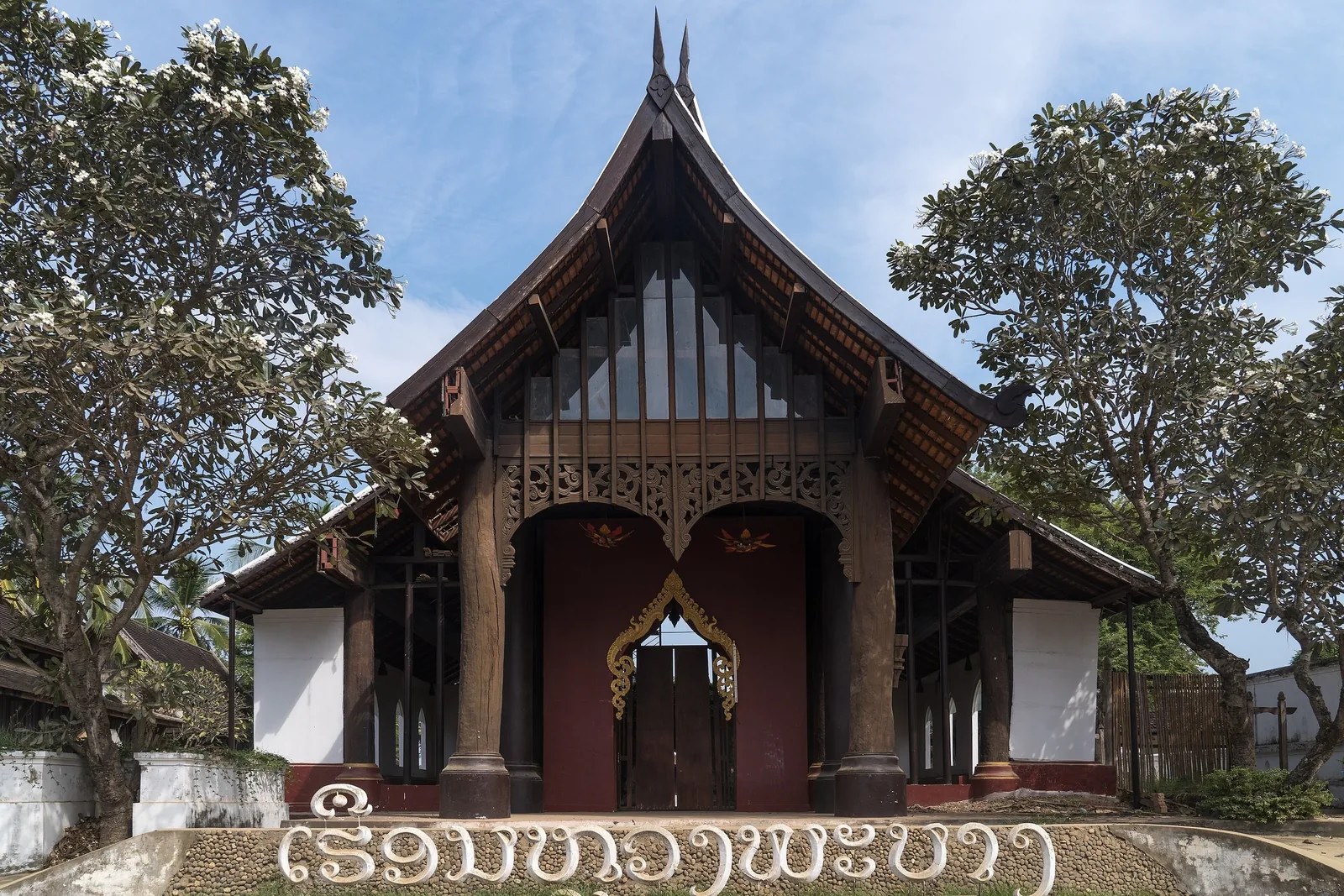 A wooden temple with a steep carved roof stands between blossoming trees in Luang Prabang.